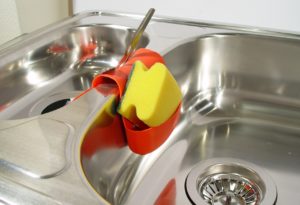 Eine silberne Küchenspüle mit zwei Spülbecken. In der Mitte der zwei Becken hängt darüber ein rotes Aufbewahrungsfach mit einem gelben Schwamm und einem silbernen Löffel darin.