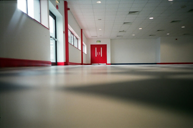 Ein großer Raum mit einem ganz ebenen, grauen Boden, am hinteren Ende sieht man eine rote Tür.