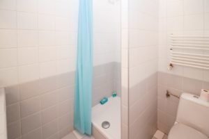 Man sieht ein gefliestes Badezimmer mit einer Dusche, einer Toilette und einer Handtuchheizung. Vor der Dusche hängt ein hellblauer Duschvorhangn und in der Dusche stehen hellblaue Utensilien farblich passend. Auf dem Toilettenspülkasten liegt eine Rolle Klopapier. Die Fließen sind im unteren Bereich des Badezimmers grau und im oberen Bereich weiß.