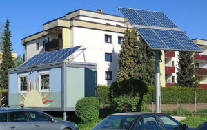 Auf einem bunten Container und an einer Straße sind Solarkollektoren aufgestellt. Im Vordergrund sind zwei Autos zu sehen, im Hintergund ein weißes, modernes Haus, sowie rote Balkone.