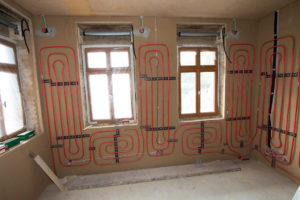 Ein Raum im Rohbau mit drei Fenstern. An der Wand wurden die roten Leitungen einer Wandheizung verlegt.