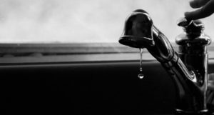 Man erkennt auf diesem schwarz-weiß Foto einen tropfenden Wasserhahn. Ein Finger drückt den Wasserhebel nach unten.