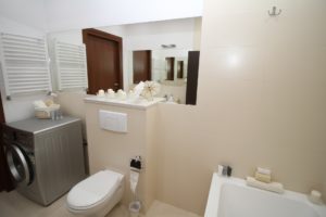 Ein in beige-weiß gehaltenes Badezimmer mit Blick direkt auf die weiße Toilette mit Unterputz WC-Spülkasten, den großen Wandspiegel sowie die silber-graue Waschmaschine. Es sieht edel aus und beinhaltet kleinere Design-Utensilien.