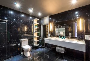 Man sieht ein dunkel gefliestes großes Badezimmer mit einem großen Spiegel und zwei Waschbecken. An der Decke gibt es Leuchtspots und links und rechts neben dem Spiegel sind weitere Badezimmerleuchten installiert.
