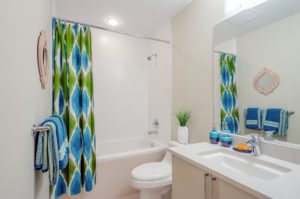 Ein Badezimmer mit weißen Wänden und einem grün-blauen Duschvorhang, der farblich heraussticht. Man sieht in dem Badezimmer ein Waschbecken, eine Toilette und eine Badewanne, wo der Vorhang seitlich davor hängt.
