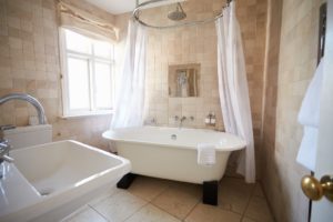 Eine freistehende Badewanne in einem beige gefliesten Badezimmer. Über der Badewanne hängt eine Duschvorhanghalterung mit einem weißen Vorhang. Es sieht alles edel und rustikal zugleich aus.