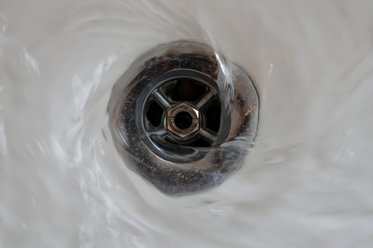 Ein silberner Waschbeckenabfluss eingefasst in weißer Keramik. Wasser läuft in einem Strudel ab.
