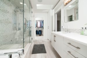 Es ist ein weiß gehaltenes Badezimmer zu sehen, durch welches man direkt in einen Kleiderschrank mit Hemden und Hosen schauen kann. Links vor der großen, mit weiß-grauen Mosaiksteinen verzierten Dusche liegt ein grauer Badteppich. Es wirkt alles sehr hell und offen.